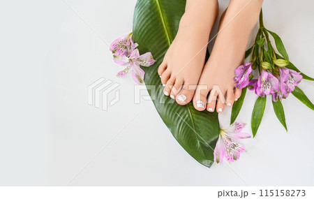 Beautiful perfect female skin legs feet top view with tropical flowers and green palm leaf. Nail polish, care and clean, spa pedicure treatment in white. Concept on background isolated. Copy space 115158273