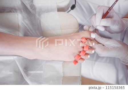 Pedicurist doing white nail polish on client legs using shellac lamp. Professional medical pedicure procedure. Foot treatment in SPA salon. Podiatry clinic. Beautician doctor hands in white gloves. 115158365