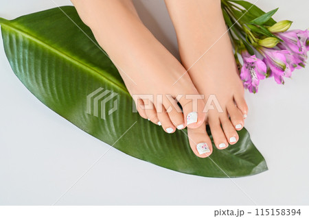 Beautiful perfect female skin legs feet top view with tropical flowers and green palm leaf. Nail polish, care and clean, spa pedicure treatment in white. Concept on background isolated. Copy space 115158394