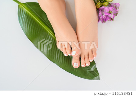 Beautiful perfect female skin legs feet top view with tropical flowers and green palm leaf. Nail polish, care and clean, spa pedicure treatment in white. Concept on background isolated. Copy space 115158451