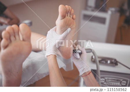 Podiatrist using grinding equipment and making procedure polish for feet pedicure. Podology beautician in white gloves cleaning skin of client legs from callus and corn with professional tool. Podiatrist using grinding equipment and making procedure polish for feet pedicure. Podology beautician in white gloves cleaning skin of client legs from callus and corn with professional tool. 115158562