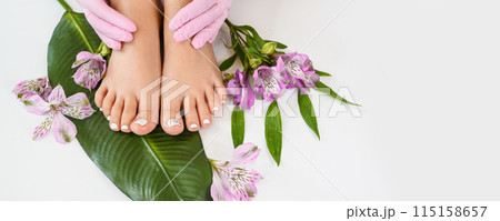 Beautiful perfect skin female legs feet top view with tropical flowers and green palm leaf. Hands of beautician in pink gloves. Nail polish, care and clean, spa pedicure treatment in white. Copy space 115158657