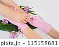 Beautiful perfect skin female legs feet top view with tropical flowers and green palm leaf. Hands of beautician in pink gloves. Nail polish, care and clean, spa pedicure treatment in white. Copy space 115158681