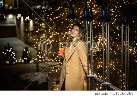 beautiful young girl in a beige coat with a paper glass of coffee against the backdrop of many bokeh lights 115166269