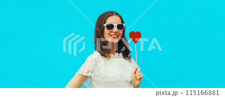 Portrait of beautiful young woman with red heart shaped lollipop on stick on blue studio background Portrait of beautiful young woman with red heart shaped lollipop on stick on blue studio background 115166881