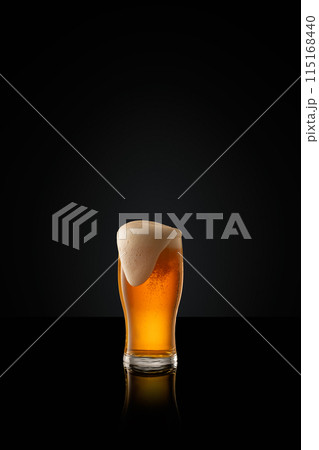 Tall glass filled with amber colored lager beer, topped with creamy white head against black studio background. Tall glass filled with amber colored lager beer, topped with creamy white head against black studio background. 115168440