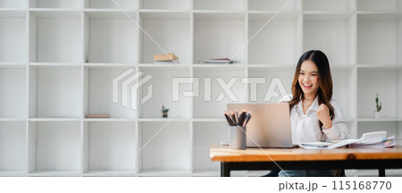 Young woman working on a laptop in a modern office, celebrating success with a smile. Minimalist bookshelf background. Young woman working on a laptop in a modern office, celebrating success with a smile. Minimalist bookshelf background. 115168770