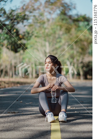 Young woman sitting on a park pathway, holding a water bottle, surrounded by trees and nature, enjoying a sunny day. Young woman sitting on a park pathway, holding a water bottle, surrounded by trees and nature, enjoying a sunny day. 115168776