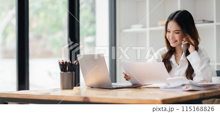 Young woman working from home office, using laptop and documents, smiling and talking on phone in bright, modern workspace. 115168826