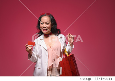 Attractive middle age woman holding credit card and shopping bags on red background 115169034