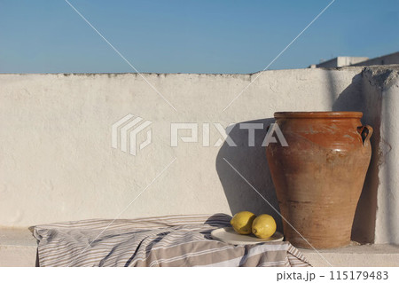 Vintage olive clay storage pot, vase with fresh lemon fruit and textile throw, blanket. White old textured white wall mockup in sunlight. Blue sky background. Summer still life, breakfast composition. 115179483