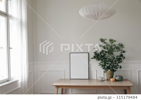 Elegant Scandinavian living room interior. Black thin frame poster, picture frame mockup on office table near window. Green tree branches in vase, pumpkins, cup of coffee. Linen lamp shade. Home decor Elegant Scandinavian living room interior. Black thin frame poster, picture frame mockup on office table near window. Green tree branches in vase, pumpkins, cup of coffee. Linen lamp shade. Home decor 115179484