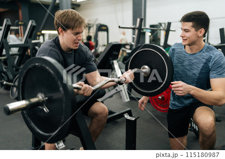 Exhausted male exercising support under supervision of personal coach. Trainer helping beginner sportsman for correct doing Scott Curls with barbell, lifting up EZ-bar sitting on preacher curl bench. 115180987
