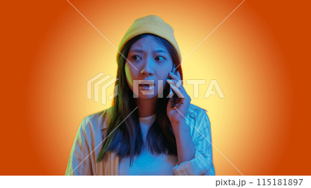 Phone scam. Mobile fraud. Blue color neon light overwhelmed hipster girl talking using gadget isolated on orange gradient copy space background. 115181897