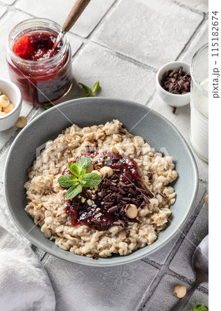 Oatmeal porridge with raspberry jam and nuts in gray bowl on gray tile background served with spoon. Healthy diet breakfast Oatmeal porridge with raspberry jam and nuts in gray bowl on gray tile background served with spoon. Healthy diet breakfast 115182674