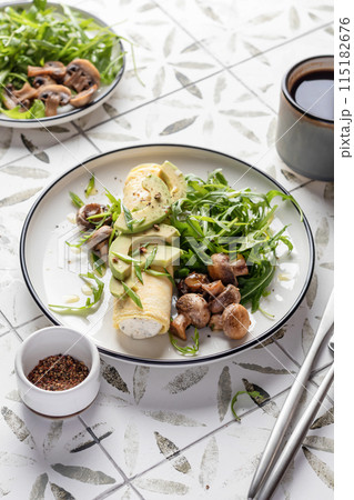 French rolled omelette stuffed with cream cheese with avocado, aragula and mushrooms for breakfast on white plate printed tile background, text space 115182676