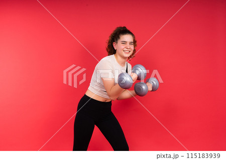 Teenage sportive girl exercises with dumbbells to develop muscles isolated on red background. 115183939