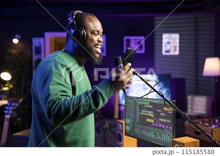 Pleased artist does a recording session in his home studio with professional gear, performing a new song on microphone. Smiling composer reading the lyrics and notes on smartphone. Pleased artist does a recording session in his home studio with professional gear, performing a new song on microphone. Smiling composer reading the lyrics and notes on smartphone. 115185580