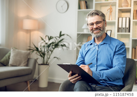 Man psychologist wearing blue shirt holding clipboard standing in his office Man psychologist wearing blue shirt holding clipboard standing in his office 115187458