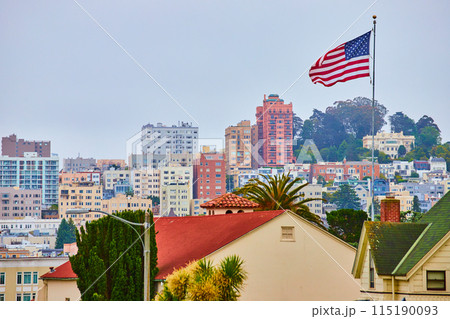 American Flag in wind over residential rooftops with overcast sky and city behind it American Flag in wind over residential rooftops with overcast sky and city behind it 115190093