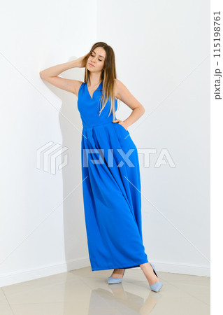 Dreamy woman in long blue sleeveless dress leaning to white wall 115198761