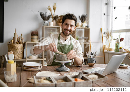 Bearded male seated at wooden table with various tools, materials, and laptop. 115202112