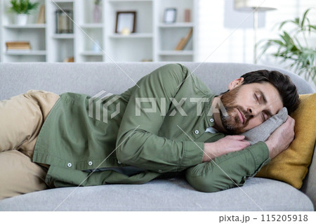 Man taking a nap on a comfortable couch in a well-lit living room. Relaxation and rest in a cozy home environment. Man taking a nap on a comfortable couch in a well-lit living room. Relaxation and rest in a cozy home environment. 115205918