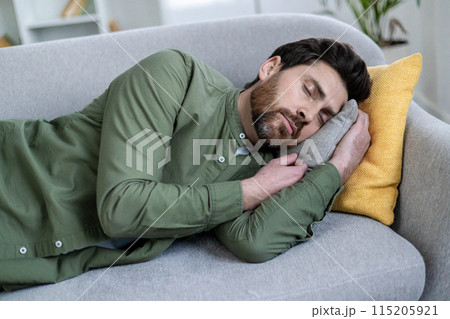 Bearded man peacefully napping on a gray sofa with yellow and gray pillows in a cozy living room environment. Bearded man peacefully napping on a gray sofa with yellow and gray pillows in a cozy living room environment. 115205921