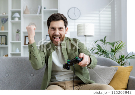 Bearded man enthusiastically playing video games on a couch in a stylish, well-lit living room. He is celebrating a victory while holding a game controller. Bearded man enthusiastically playing video games on a couch in a stylish, well-lit living room. He is celebrating a victory while holding a game controller. 115205942