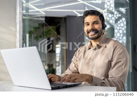 Satisfied muslim male in headset with micro using contemporary gadget by white desktop at workspace. Smiling bearded guy in beige shirt typing on keyboard of pc and smiling widely at camera. 115206040