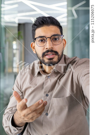 First person view of brunette man in cotton shirt wearing stylish eyeglasses and gesturing to camera during video meeting. Arab company boss sharing important information with employers of department. 115206075