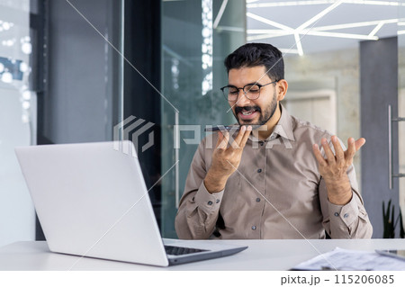 A professional man engaging with a voice assistant on his smartphone, smiling as he communicates, in a modern office setting with a laptop in front. 115206085
