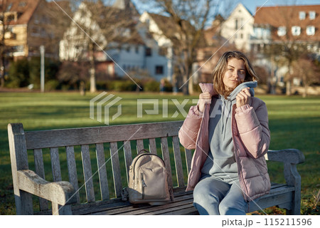 Winter Joy in Bitigheim-Bissingen: Beautiful Girl in Pink Jacket Sitting Amidst Half-Timbered Charm. beautiful girl in a pink winter jacket sitting on a bench in a park, set against the backdrop of 115211596