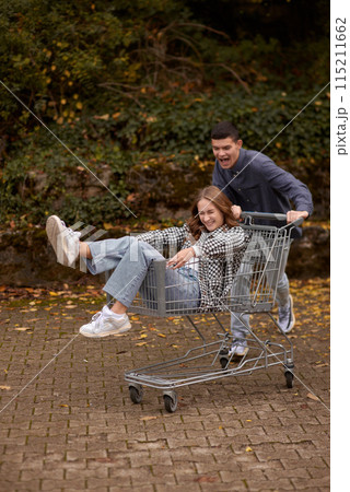 Autumn Joyride: Boy Rolling Girl in Shopping Cart amid Nature's Beauty. Teen Love's Autumn Frolic: Boy Rolling Girlfriend in a Shopping Cart 115211662