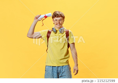 Portrait of redhaired smiling young man in glasses holding rolled-up diploma against vibrant yellow background. Portrait of redhaired smiling young man in glasses holding rolled-up diploma against vibrant yellow background. 115212599
