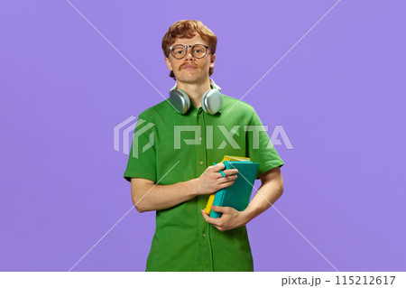Young man, with headphones on neck, dressed casual attire holding books notebooks and looks at camera confidently against purple background. Young man, with headphones on neck, dressed casual attire holding books notebooks and looks at camera confidently against purple background. 115212617
