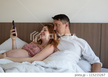 A happy couple relaxing in bed, sharing a joyful moment and looking at a smartphone together A happy couple relaxing in bed, sharing a joyful moment and looking at a smartphone together 115215267