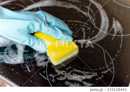 A gloved hand scrubs a black electric stovetop with a soapy sponge 115216443