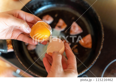 A hand cracks an egg over a frying pan of sizzling sausage slices 115216462