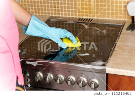 A person wearing a blue household glove cleans an electric stovetop with a yellow sponge 115216463