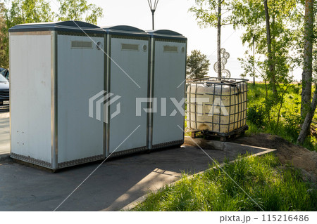 Three white portable toilets stand on a paved area in a park setting Three white portable toilets stand on a paved area in a park setting 115216486