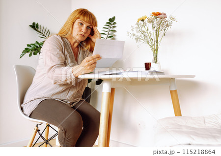 blonde woman reading letter notice while sitting at table in room blonde woman reading letter notice while sitting at table in room 115218864