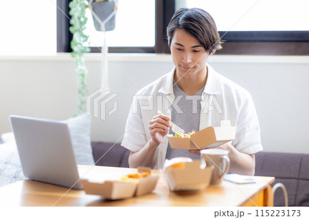 パソコンを見ながらデリバリーで注文した料理を食べる男性 パソコンを見ながらデリバリーで注文した料理を食べる男性 115223173