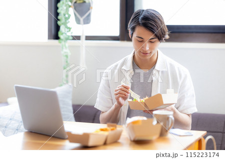 パソコンを見ながらデリバリーで注文した料理を食べる男性 パソコンを見ながらデリバリーで注文した料理を食べる男性 115223174