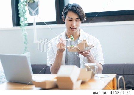 パソコンを見ながらデリバリーで注文した料理を食べる男性 パソコンを見ながらデリバリーで注文した料理を食べる男性 115223176