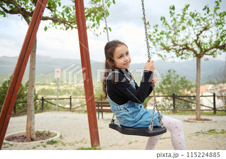 Little child girl smiling and dreamily looking away while sitting on a wooden swing in the playground outdoors. People. Happy carefree childhood. Recreation. Kids development and entertainment 115224885