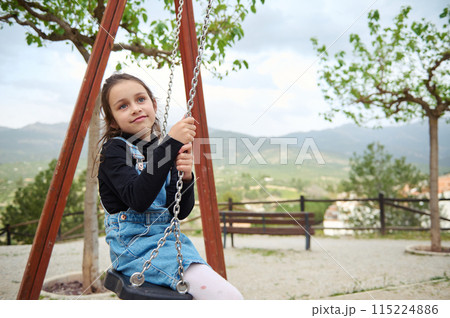 Little child girl 6 years old smiling and dreamily looking away while sitting on a wooden swing in the playground outdoors. People. Happy carefree childhood. Recreation Little child girl 6 years old smiling and dreamily looking away while sitting on a wooden swing in the playground outdoors. People. Happy carefree childhood. Recreation 115224886