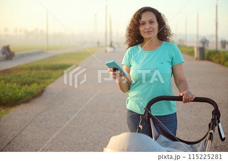 Beautiful young happy mother holding using a smart mobile phone, pushing baby pram on the promenade at sunset nd smiling looking at camera. People. Maternity leave lifestyle. Online communication Beautiful young happy mother holding using a smart mobile phone, pushing baby pram on the promenade at sunset nd smiling looking at camera. People. Maternity leave lifestyle. Online communication 115225281