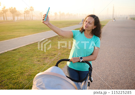 Smiling woman recording video or taking selfie while pushing baby pram on the promenade at sunset. Happy mother enjoying walk with her baby in stroller on a summer day. People. Maternity and infancy Smiling woman recording video or taking selfie while pushing baby pram on the promenade at sunset. Happy mother enjoying walk with her baby in stroller on a summer day. People. Maternity and infancy 115225285