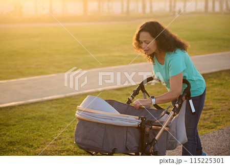 Smiling happy young woman, loving caring mother walking with her newborn baby in stroller, spending time funny on the city park t sunset Smiling happy young woman, loving caring mother walking with her newborn baby in stroller, spending time funny on the city park t sunset 115225301